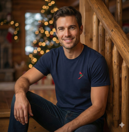 Man wearing a t-shirt with an embroidered skier on the left chest is sitting on a wooden staircase with a Christmas tree in the background